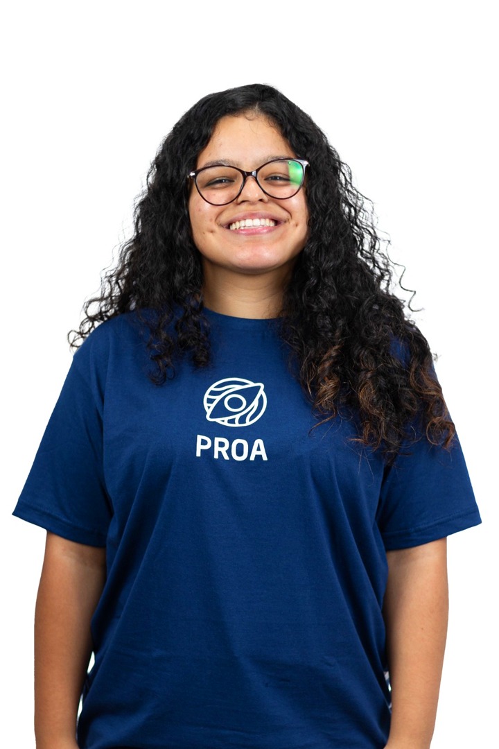 A imagem destaca uma jovem usando uma camiseta com a palavra PROA estampada nela. Ela é o ponto central da imagem e está posicionada no centro. Seu rosto é visível e ela está sorrindo para a câmera. A mulher também está usando óculos, o que realça ainda mais sua aparência. O texto PROA está localizado na parte inferior da camiseta, ligeiramente à direita. No geral, a imagem captura uma mulher alegre exibindo orgulhosamente sua camiseta com a palavra PROA.