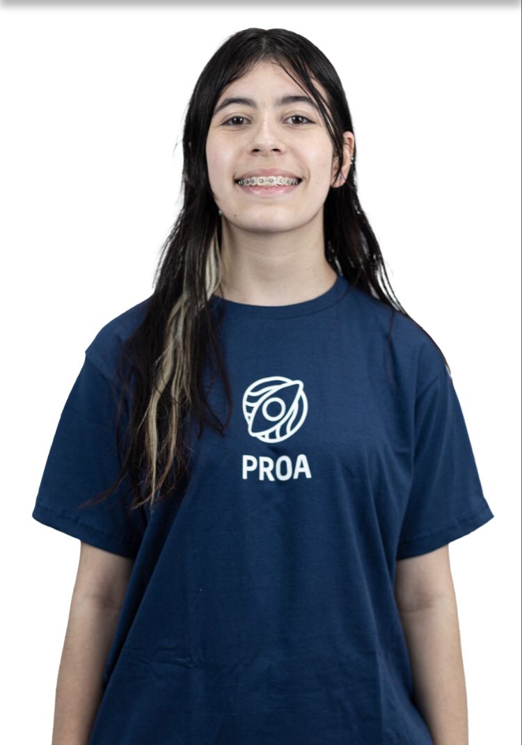 A imagem mostra uma jovem usando uma camiseta com a palavra PROA escrita nela. A camiseta é o principal ponto focal da imagem, e a palavra está exibida proeminentemente no centro da camiseta. A garota está em pé contra um fundo simples, permitindo que o texto em sua camiseta se destaque.